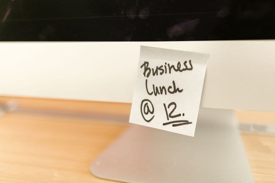 Un foglietto adesivo bianco attaccato al bordo inferiore di un monitor, scritto a mano con pennarello nero Business Lunch @ 12 e sottolineato due volte, su una scrivania di legno chiaro