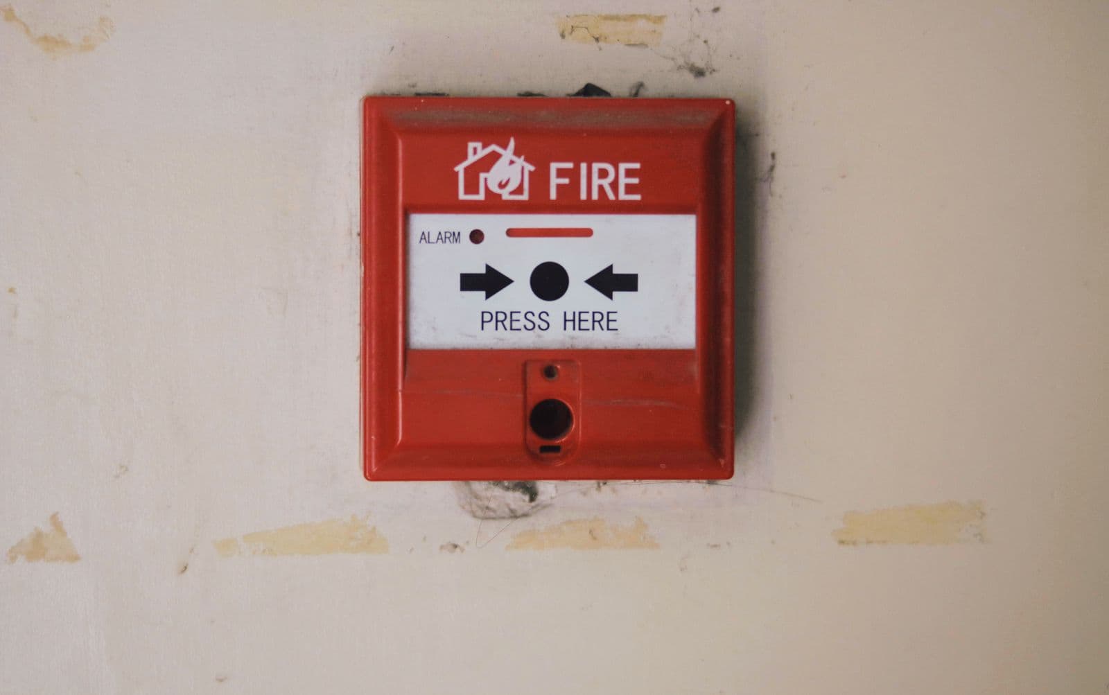 A red fire alarm call point on a beige wall, marked FIRE and PRESS HERE with two arrows pointing to the button