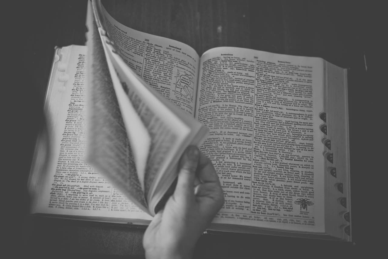 A hand turning the pages of a thick printed dictionary, thumb tabs visible along the edge, in black and white
