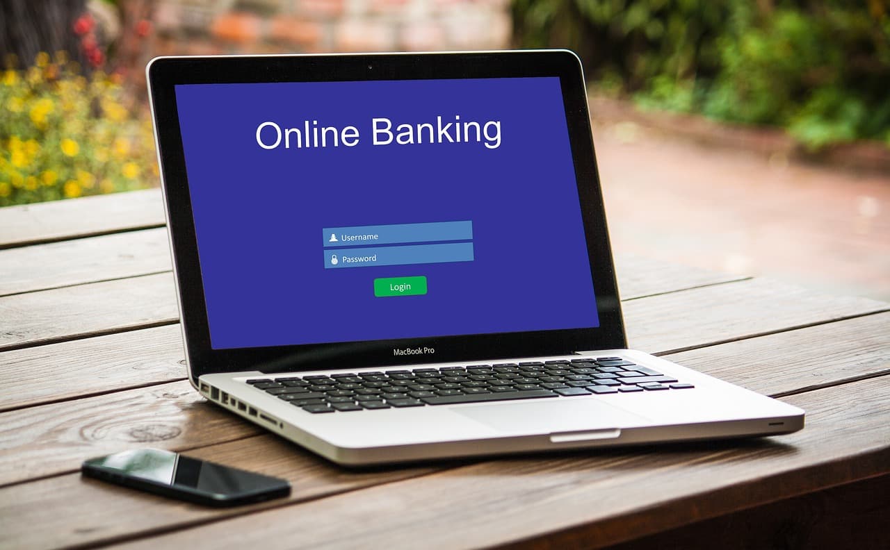 Un portatile su un tavolo di legno mostra una schermata di accesso al banking online con i campi nome utente e password, accanto a uno smartphone. I dati di contatto trapelati rendono credibile un phishing a tema login, il vero rischio qui.