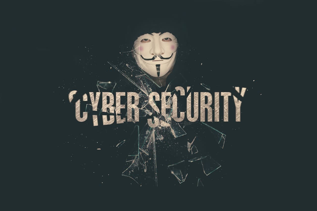 Uma pessoa a usar uma máscara de Guy Fawkes atrás de vidro partido, com as palavras 'cibersegurança'