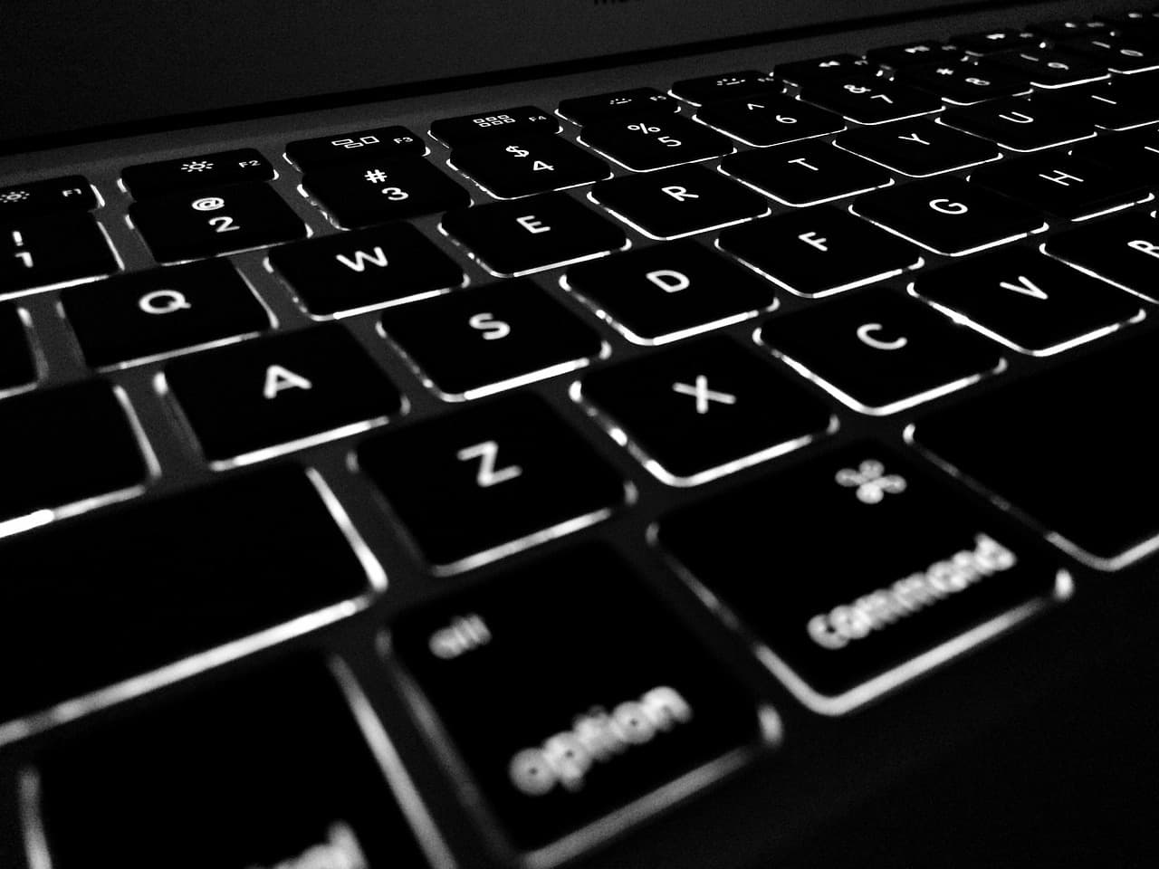A backlit laptop keyboard photographed close up in black and white, the number keys showing the at sign, hash, dollar and percent symbols above the digits.